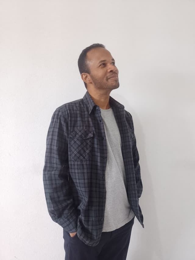 Foto do Desenvolvedor Full Stack, Rafael Lopes de perfil e olhando para cima. Ele está usando camiseta cinza clara, camisa xadrez com tons de verde escuro e preto e calça preta. O Desenvolvedor Rafael Lopes é um homem negro de cabelos e olhos pretos